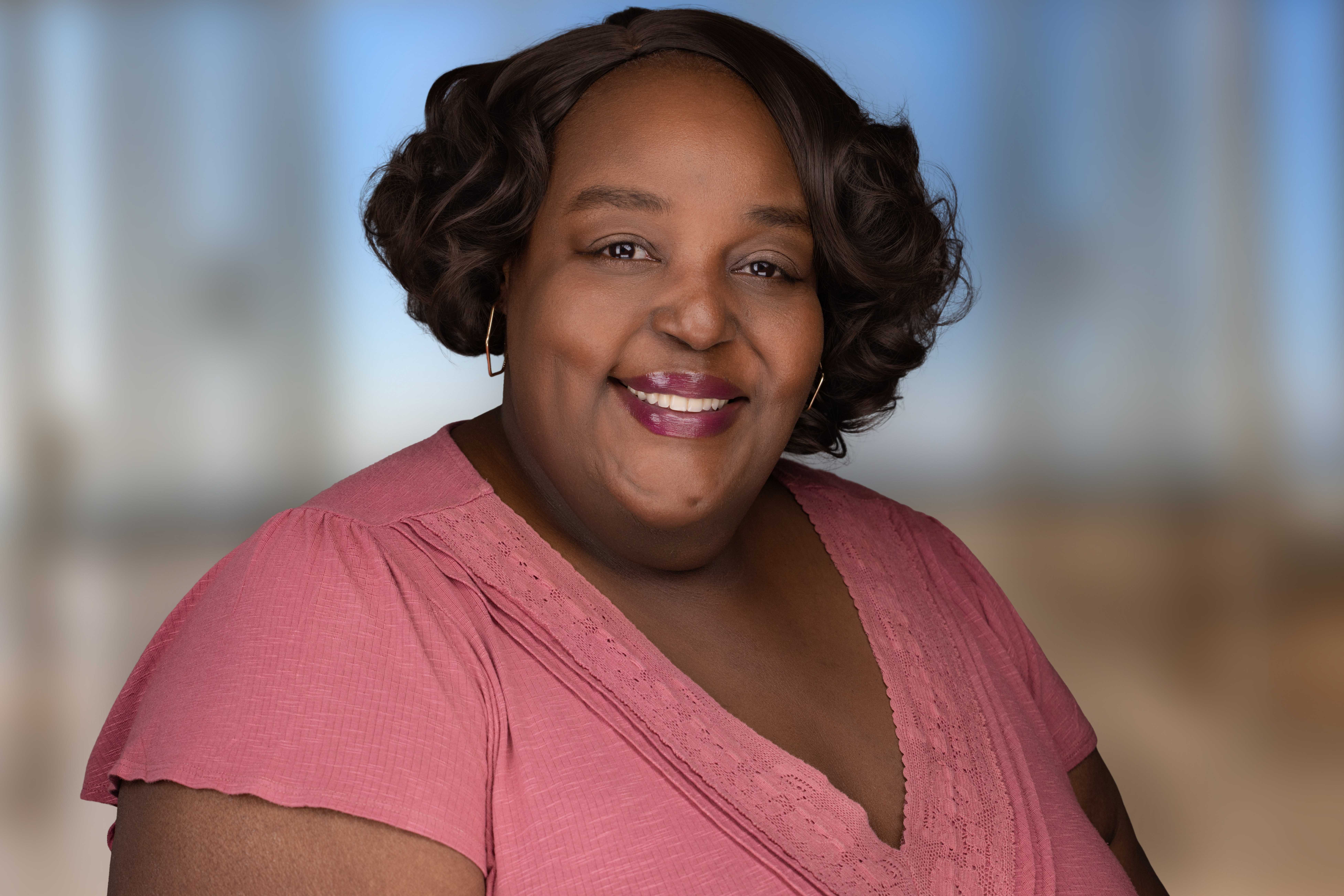 Landscape portrait of Kesha Moore smiling in a pink blouse against a softly blurred neutral office background.